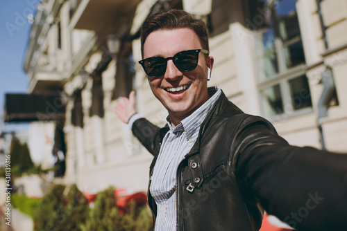 Close up young smiling fun happy caucasian man 20s in black leather jacket glasses doing selfie shot on mobile cell phone camera show downtown buildings, Concept of people urban lifestyle excursion.