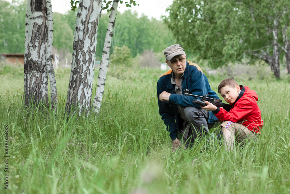 A boy in a red windbreaker is aiming an air gun with a grandfather in a cap in summer park. Concept weapons and children, respect, caution, training. A boy in a red windbreaker is aiming an air gun with a grandfather in a cap in summer park. Concept weapons and children, respect, caution, training.