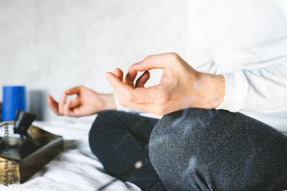 Man meditating in lotus position on bed.  Focus on incense stick and smoke. Unrecognizable yoga practitioner in the background. Relax after Yoga training