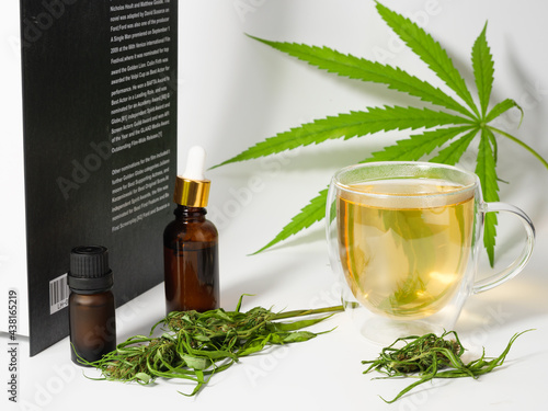 Cannabis tea cup with marijuana fresh green leaves and flower in glass cup, CBD oil brown bottle and book on white background.