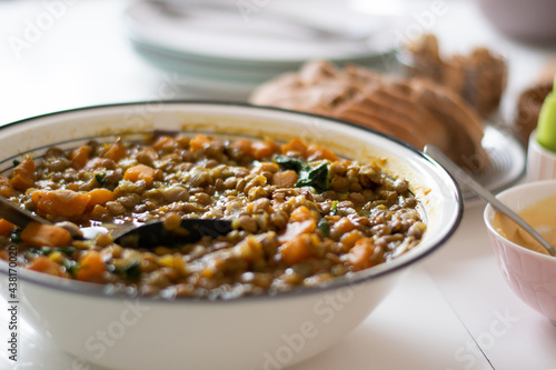 Lentils prepared in the traditional way, in a white bowl with greens, carrots and bay leaf. In the background is sliced bread as well as plates set up for use. The hummus is in a small bowl next to it
