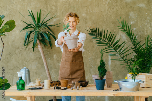 Amazing blond female flower shop owner in apron showing you pot with small green plant and smiling at camera on green background around plants in her home garden. Planting and profession concept. photo