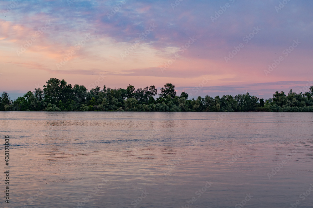 Fantastic pink sunset over the Danube river. Summer landscape with the reflection of the sky in the river, green grass and forest on the high riverbank