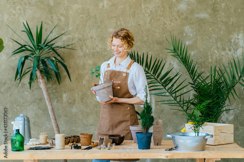 Blond housewife admires her flowers. A woman in apron looks after home flowers on green background. Flower seller. Floriculture business.