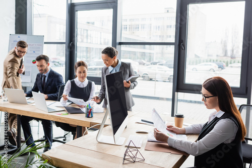 Businesswoman holding papers near coffee, computer and multiethnic colleagues in office