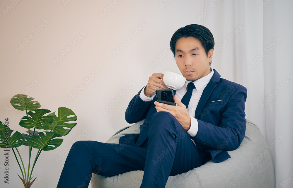 Asian Financial Advisor Drink Coffee See Smartphone on Bean Bag in Home Office. Businessman Work at Home for Social Distance or Home Quarantine in Covid 19 or Coronavirus Situation in Vintage Tone Asian Financial Advisor Drink Coffee See Smartphone on Bean Bag in Home Office. Businessman Work at Home for Social Distance or Home Quarantine in Covid 19 or Coronavirus Situation in Vintage Tone