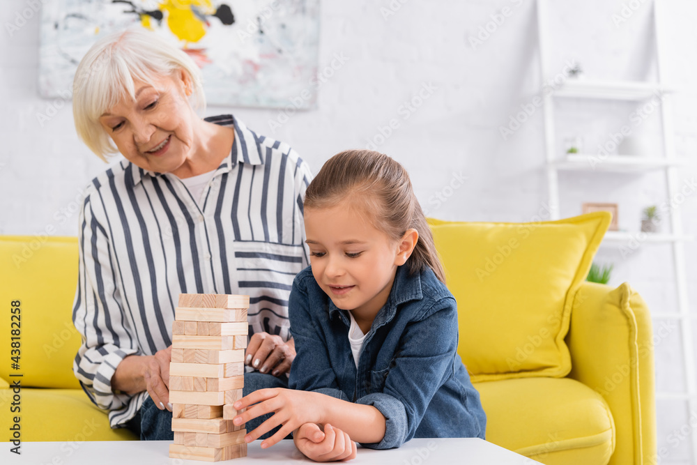 Smiling girl playing blocks wood game near blurred granny Smiling girl playing blocks wood game near blurred granny