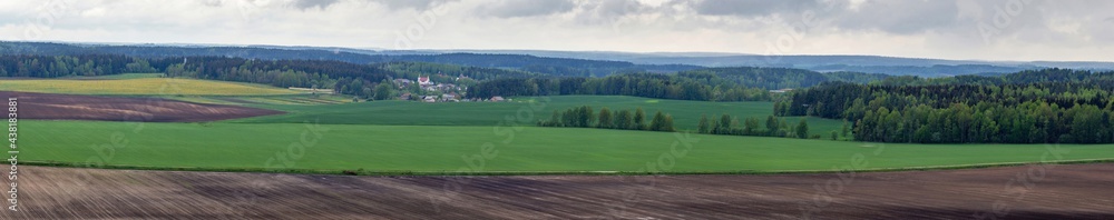 Panorama of plowed and fresh green agricultural fields under a cloudy sky. Village and forest to the horizon on the background. Detailed natural landscape with fields and forests of Belarus