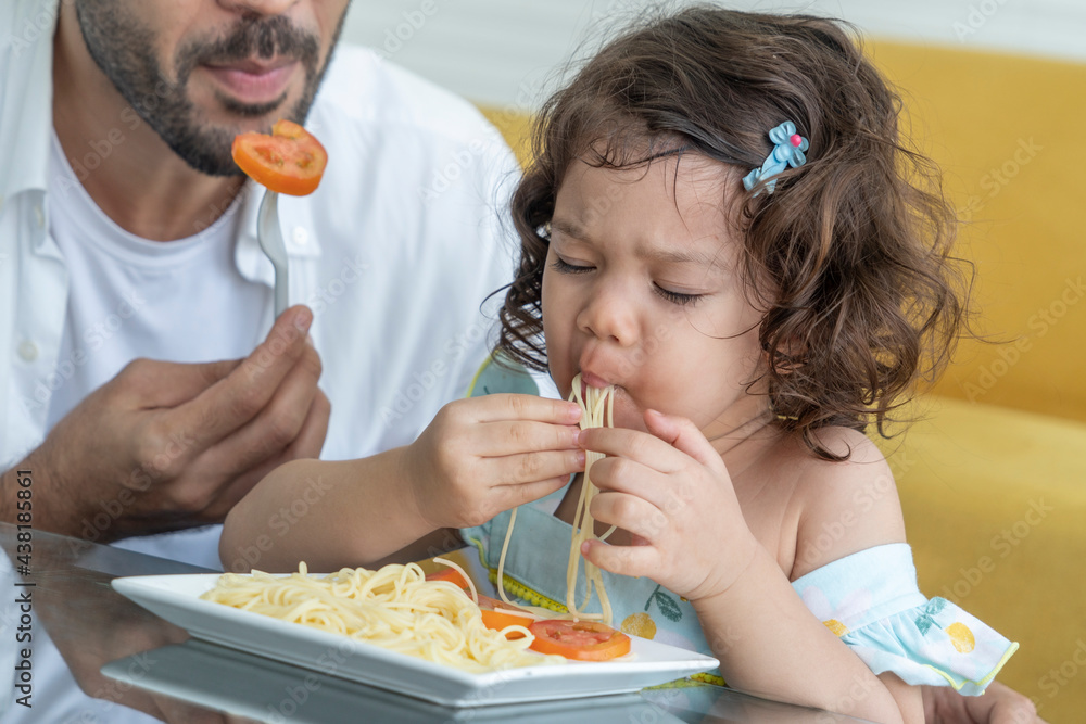 Hungry little daughter sits and eats spaghetti noodles with a spoon and eats deliciously in the kitchen at home. Hungry little daughter sits and eats spaghetti noodles with a spoon and eats deliciously in the kitchen at home.