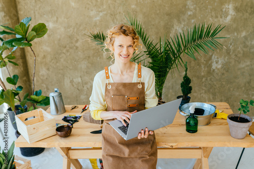 Happy blond woman gardener taking care of houseplants housewife using laptop relaxing leaning on wooden table in modern cozy interior, over green wall. photo