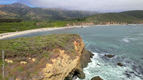 Aerial drone flying alongside ocean cliffs with greenery and mountains in the background.
