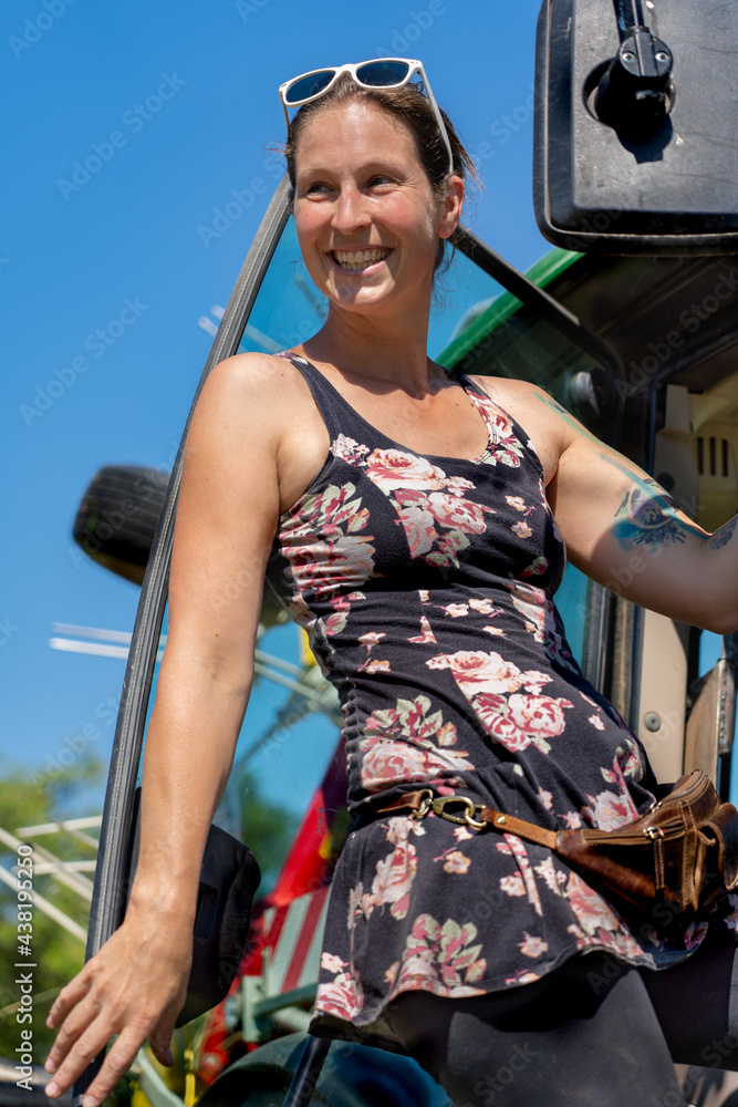 Low angle view of beautiful happy young woman upstairs on her tractor at sunny day. Vertical photo.