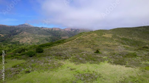 Aerial drone flying alongside ocean cliffs with greenery and mountains in the background.