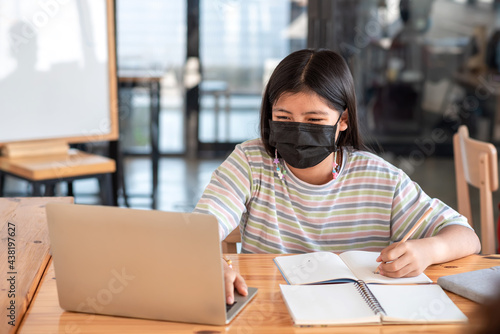 Asian woman sitting in room using laptop and taking notes wearing a mask to prevent germs.