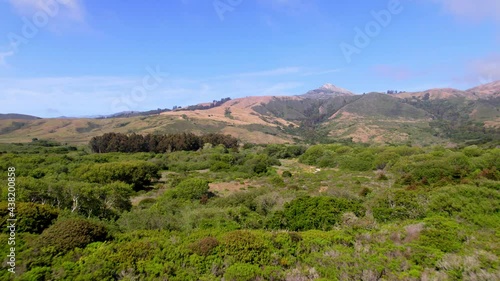 Aerial drone flying alongside ocean cliffs with greenery and mountains in the background.