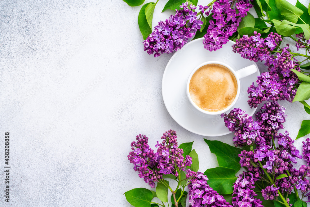 Morning cup of cappuccino with foam and branch of blooming purple lilac with green leaves on textured grey background. Coffee break. Slow life concept. Spring flowers. Flat lay, top view, copy space. Morning cup of cappuccino with foam and branch of blooming purple lilac with green leaves on textured grey background. Coffee break. Slow life concept. Spring flowers. Flat lay, top view, copy space.