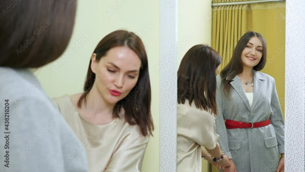 Professional sales assistant helps young woman in new grey jacket standing near large mirror in department store fitting room closeup Professional sales assistant helps young woman in new grey jacket standing near large mirror in department store fitting room closeup