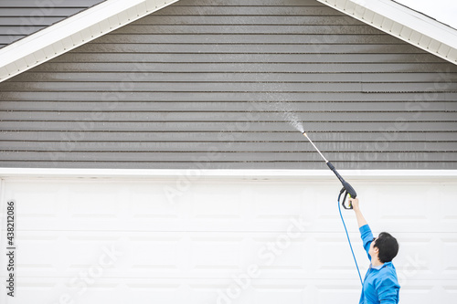 Man in blue jacket clean dusk and dirt from exterior siding under the roof at front door garage by high pressure nozzle spray with water soap cleaner, weekend chore. Washer machine of cleansing house.