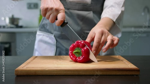 Female hands cut red bell pepper into parts by knife on wooden board in kitchen