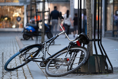 Billede på lærred Black bicycle is half on the ground and half against a tree in a shopping street, parked carelessly or overturned
