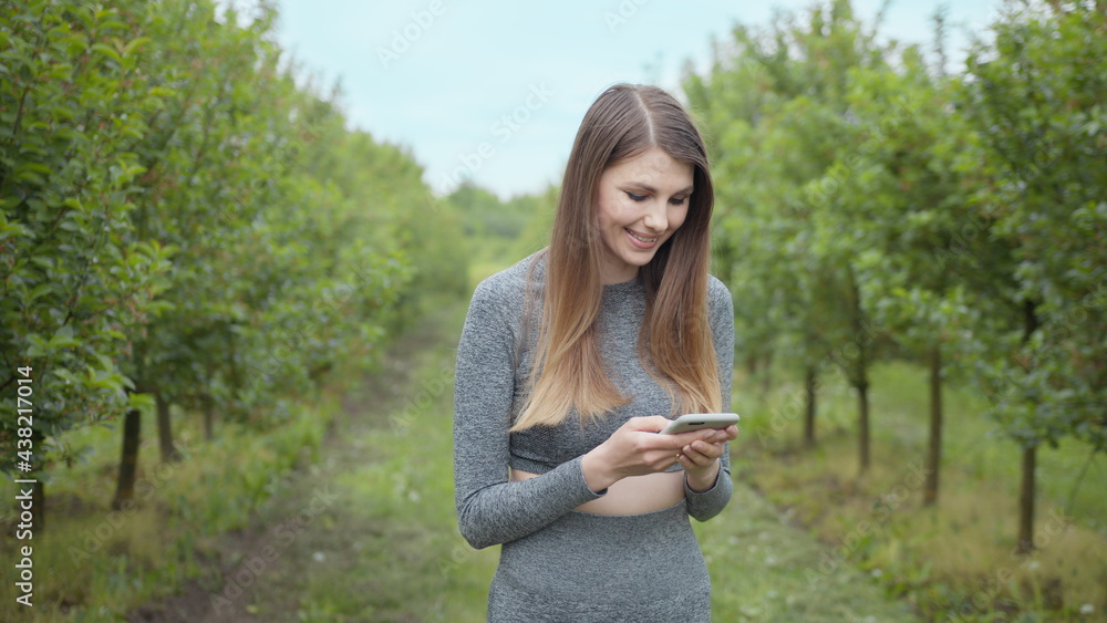 Happy woman typing by mobile phone outdoors. Cheerful girl with smartphone in park on a background. Smiling lady holding cellphone in hands outside. Athletic fitness girl holding a phone in her hands. Happy woman typing by mobile phone outdoors. Cheerful girl with smartphone in park on a background. Smiling lady holding cellphone in hands outside. Athletic fitness girl holding a phone in her hands.