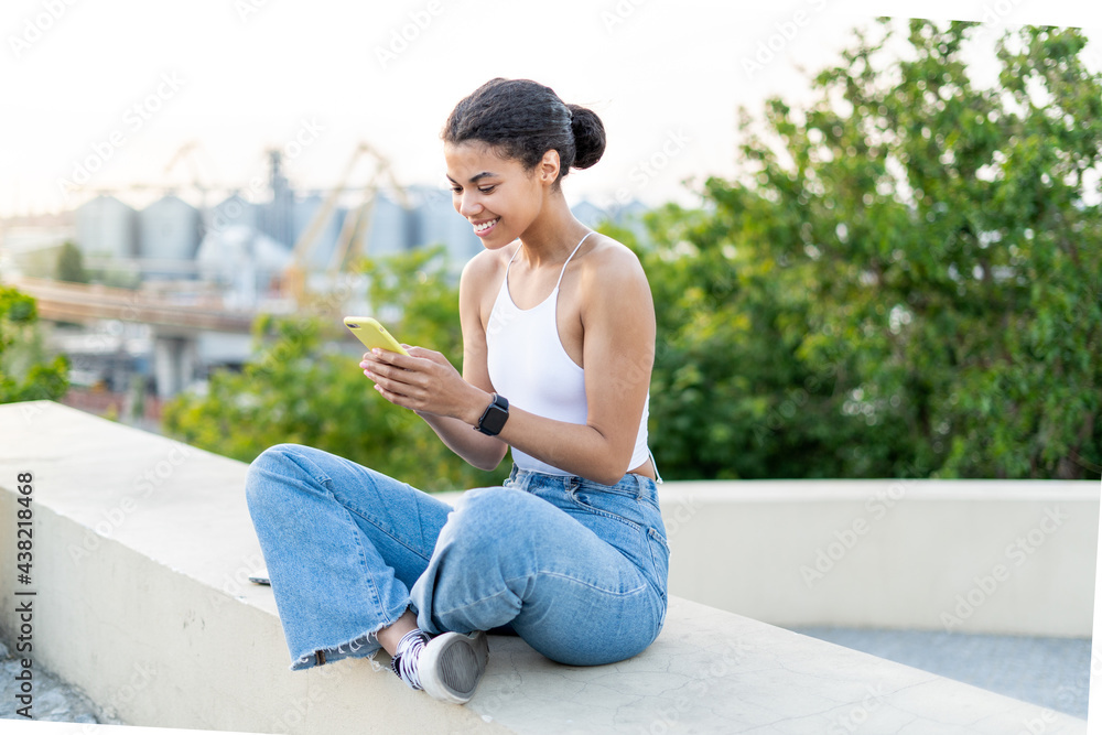 Young smily African American woman texting on her mobile phone sitting outside with a great view