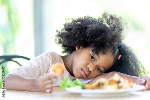 A young, curly haired African American girl sits looking at food. Childhood concepts and healthy eating.