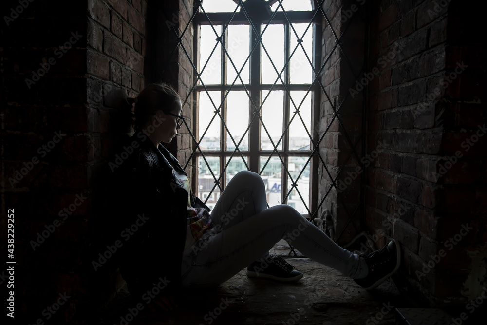 Silhouette of a girl sitting near the window. Ancient room with a window. Abandoned house. Silhouette of a beautiful girl. Light in the window.