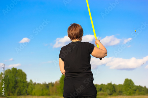 Fisherwoman with a yellow fishing rod on a background of a blue sky and green trees on a summer day. Woman catching fish. Active leisure and recreation in nature reserve. Angling on a bank of a river.