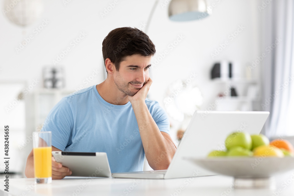 Indoor shot of a smiling European young man surfing the internet on a laptop, messaging on via social media while having a fresh breakfast at home at the kitchen table. Indoor shot of a smiling European young man surfing the internet on a laptop, messaging on via social media while having a fresh breakfast at home at the kitchen table.