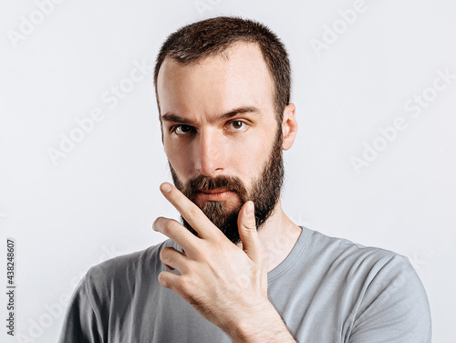 Serious puzzled handsome male model with beard holding hand on chin as if thinking about something, squinting at camera with suspicious look and standing over gray background. Man decides what to buy photo