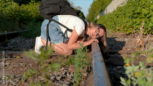 A woman with a backpack on her back puts her ear to the railway tracks. Travel on railroad ties. Old train track