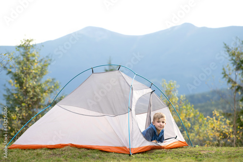 Happy child boy resting alone in a tourist tent at mountain campsite enjoying view of beautiful summer nature. Hiking and active way of life concept.