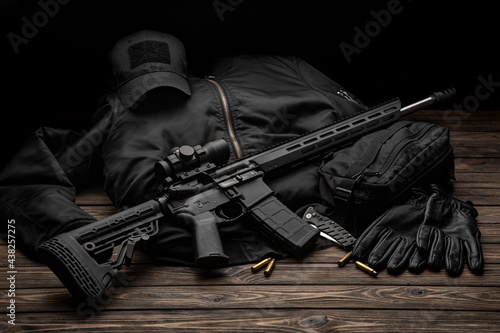 Modern automatic rifle with a telescopic sight on a dark background. Jacket, cap, gloves on a wooden table. The uniform of a guard or a mercenary