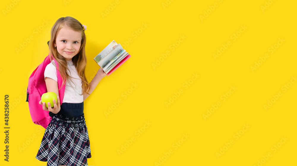 Back to school. Little blonde girl with school backpack. An elementary school student smiles and holds book and green apple on yellow background. Back to school. Little blonde girl with school backpack. An elementary school student smiles and holds book and green apple on yellow background.