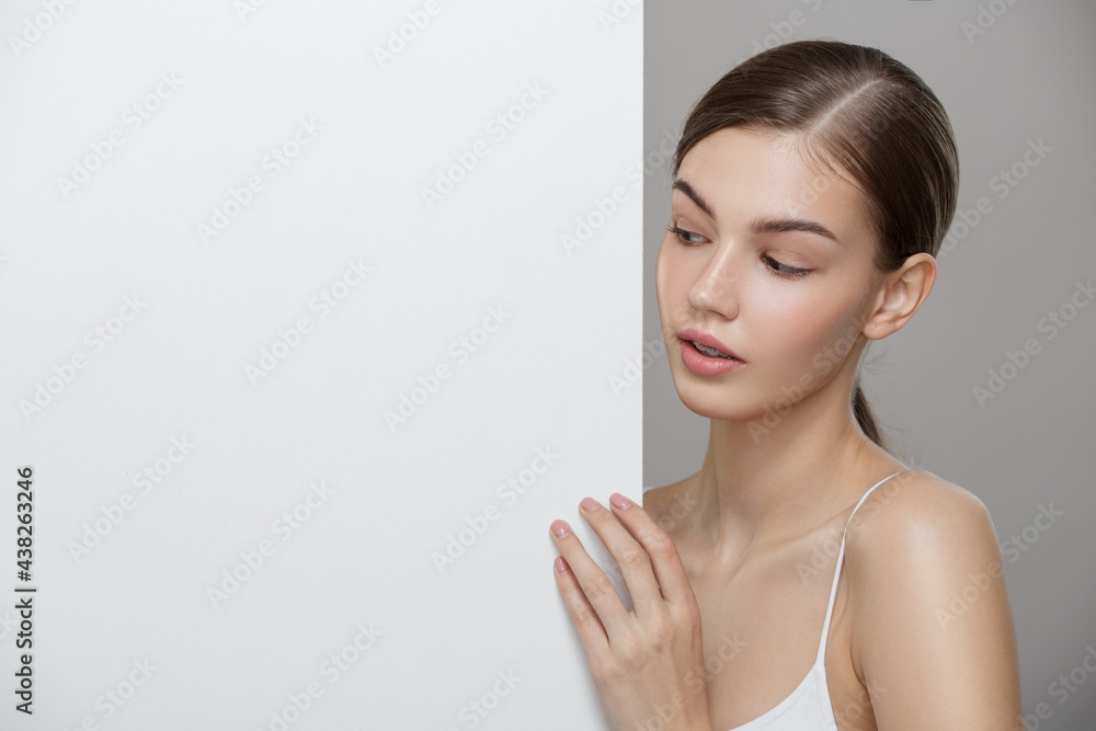 Beauty portrait of a young beautiful girl with clean skin and white empty board. Gray background. Beauty portrait of a young beautiful girl with clean skin and white empty board. Gray background.