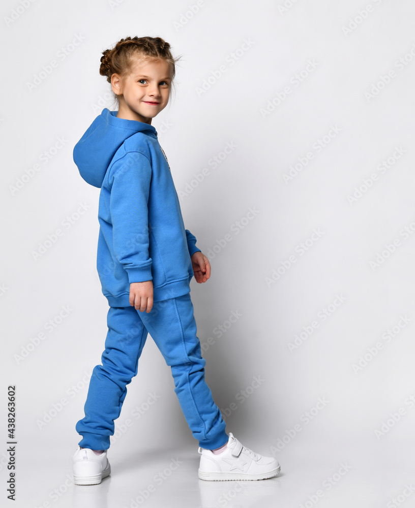 Active playful little girl in a trendy blue tracksuit having fun on a white background. A child with an emotional expression is standing half-sided and posing. Promotion of children's sports fashion. Active playful little girl in a trendy blue tracksuit having fun on a white background. A child with an emotional expression is standing half-sided and posing. Promotion of children's sports fashion.
