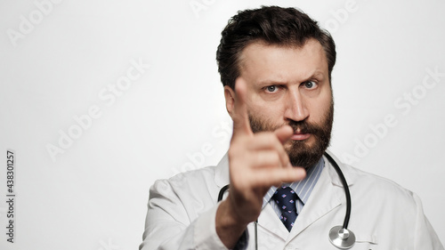 Doctor threatens with his finger. Serious frowning man doctor on white background looking at camera and points menacingly with his index finger