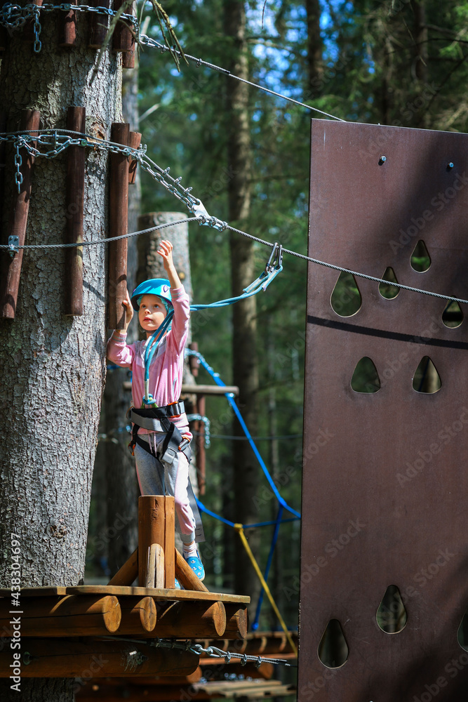 The child is engaged in sports.A girl overcomes sports obstacles in a rope park.A little girl in a protective helmet and tracksuit.Active recreation in the summer.Sports Rope Park for children