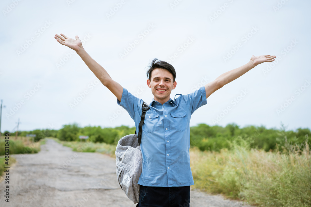 Happy young man posing with raised arms, standing on the road and looking into the distance. Happy guy in a blue shirt with a backpack. Free student enjoys vacation