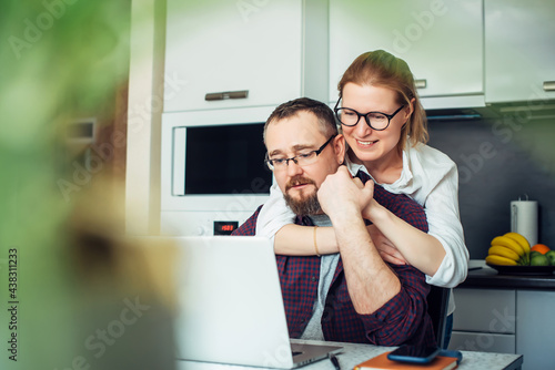 Adult married couple in cozy home interior. Husband and wife look at the laptop screen, discussing family budget. Woman embraces man from behind. In the foreground leaves of houseplant, blurred focus.