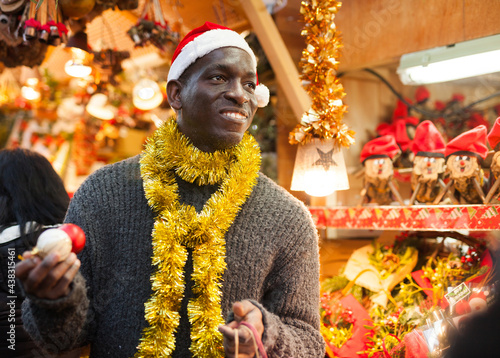 Smiling African American man in Santa hat selecting festive home decoration at outdoor Christmas fair..