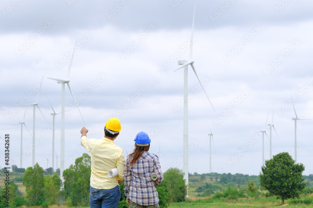 An engineer stands holding a blueprint and looks at a wind turbine project to generate electricity and check wind direction.