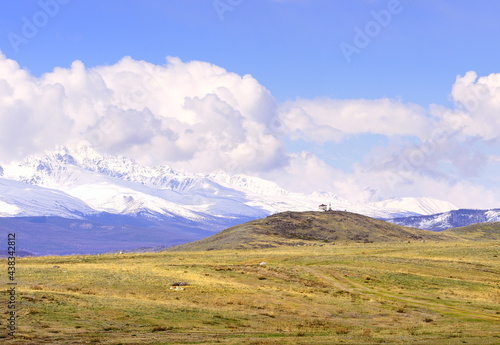 Kurai steppe in spring. Viewing gazebo on a hilltop in the middle of dry grass, snow-capped peaks of the Northern Chui range under a cloudy blue sky. Gorny Altai, Siberia, Russia