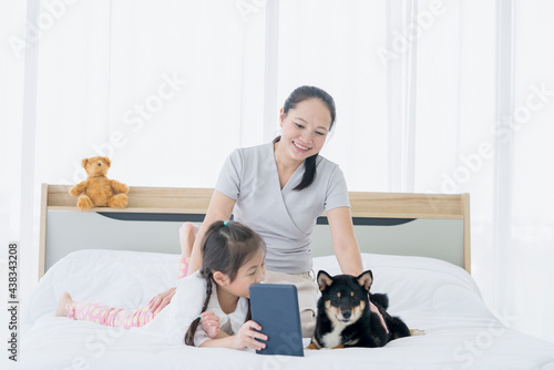 A family with a mother and daughter using a tablet in bed in the bedroom with a black Shiba Inu.
