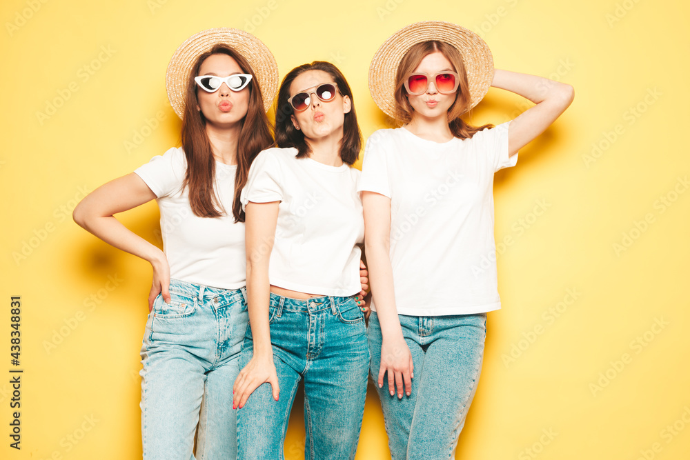 Fototapeta premium Three young beautiful smiling hipster female in trendy summer white t-shirt and jeans clothes.Sexy carefree women posing near yellow wall in studio.Positive models in hats