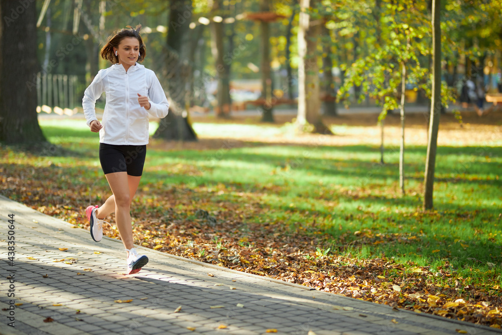 Active young woman in sportswear running at green park during morning time. Happy female with dark hair enjoying sport activity on fresh air.