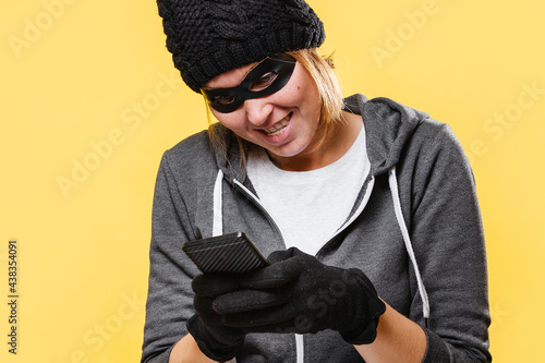 A female hacker wearing a black hat, gloves and mask holds a mobile phone in her hands, maliciously trying to hack into a database. Yellow background. The concept of cybercrime and hacking photo