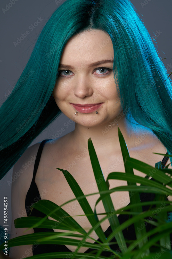 Naklejka premium Girl with blue green hair on a gray background, blue backlighting. Tropical plants around. Bright colored appearance of a creative person