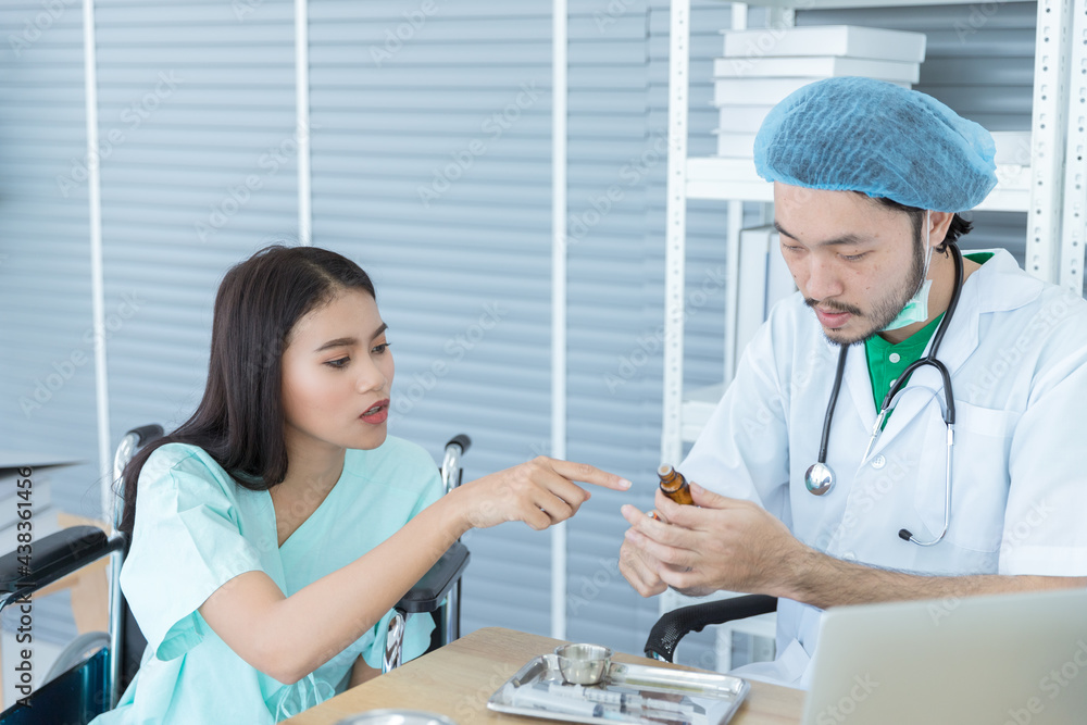 Obraz premium Smart Asian male doctor with white coat is looking at medical tablet pills while diagnosing and talking to beautiful female patient is sitting on wheelchair fort treatment in hospitsl room.